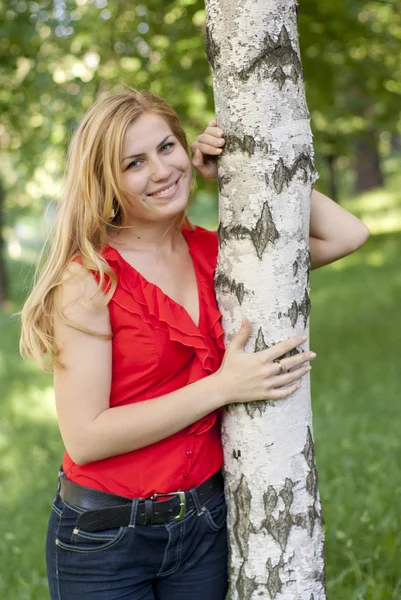 Προσωπογραφία του κοριτσιού σε μια κόκκινη μπλούζα που χαμογελάει και να αγκαλιάζει ένα σημύδα. — Φωτογραφία Αρχείου