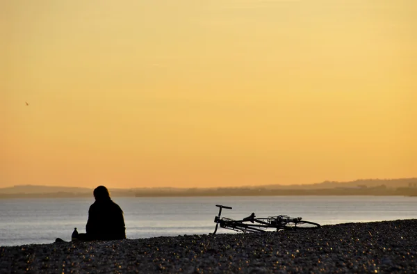 Um homem sentado e sua bicicleta em uma praia ao pôr do sol — Fotografia de Stock