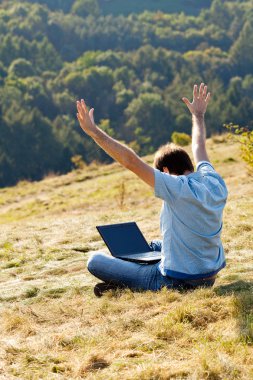 Young man using laptop sitting on the grass on the hillside clipart