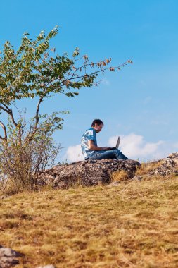 Young man using laptop sitting on a hill against the blue sky clipart