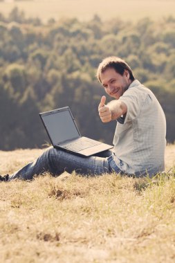 Young man using laptop sitting on the grass on the hillside clipart