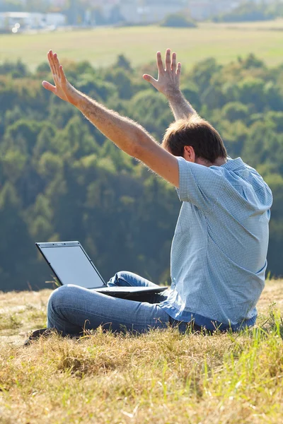Νεαρός άνδρας, χρησιμοποιώντας φορητό υπολογιστή που κάθεται στο γρασίδι στην πλαγιά του λόφου — Φωτογραφία Αρχείου