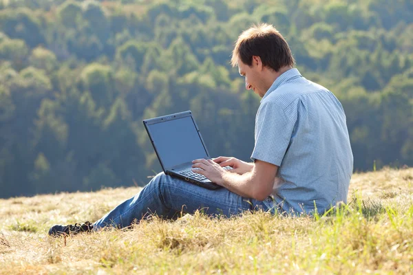 使用便携式计算机，坐在山坡上的草上的年轻人 — 图库照片