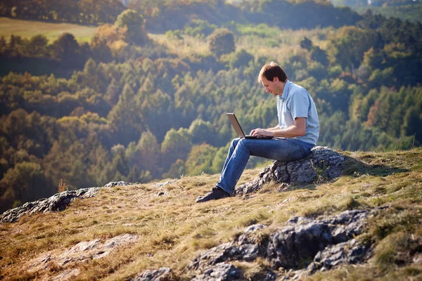 Молодий чоловік, використовуючи ноутбук, сидить на траві на схилі пагорба — стокове фото