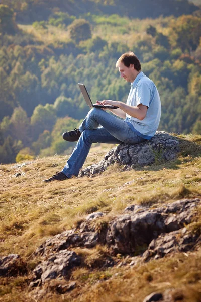 Młody człowiek za pomocą laptopa siedząc na trawie, na wzgórzu — Zdjęcie stockowe