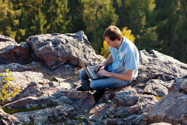 Jovem homem usando laptop sentado em uma encosta de rocha — Fotografia de Stock