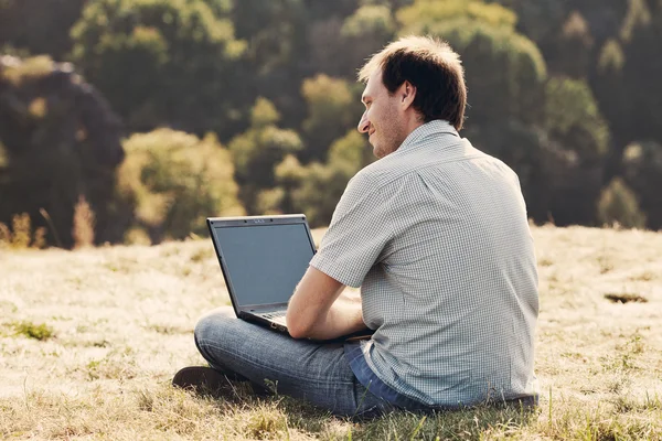 Mladý muž pomocí přenosného počítače sedět na trávě, na kopci — Stock fotografie