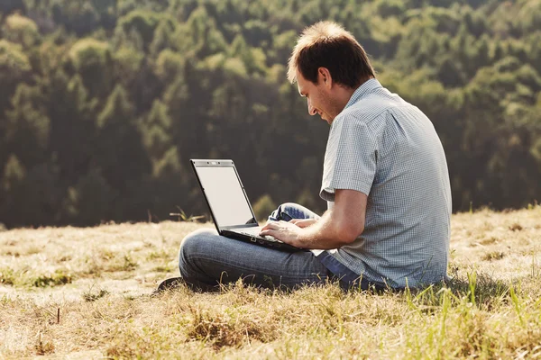 Young man using laptop sitting on the grass on the hillside — Stock Photo, Image