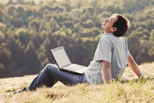 Молодий чоловік, використовуючи ноутбук, сидить на траві на схилі пагорба — стокове фото