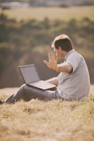 Młody człowiek za pomocą laptopa siedząc na trawie, na wzgórzu — Zdjęcie stockowe