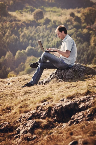 Νεαρός άνδρας, χρησιμοποιώντας φορητό υπολογιστή που κάθεται στο γρασίδι στην πλαγιά του λόφου — Φωτογραφία Αρχείου