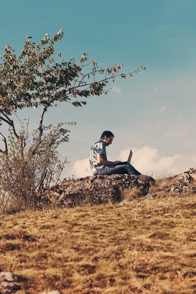 Young man using laptop sitting on a hill against the blue sky — Stock Photo, Image