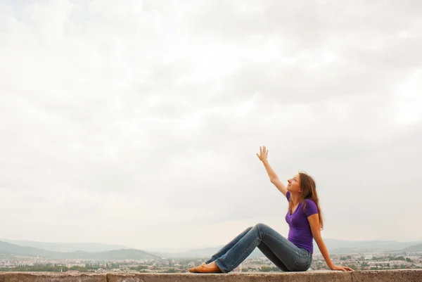 Young woman sitting with raised hand against blue sky — Stock Photo, Image