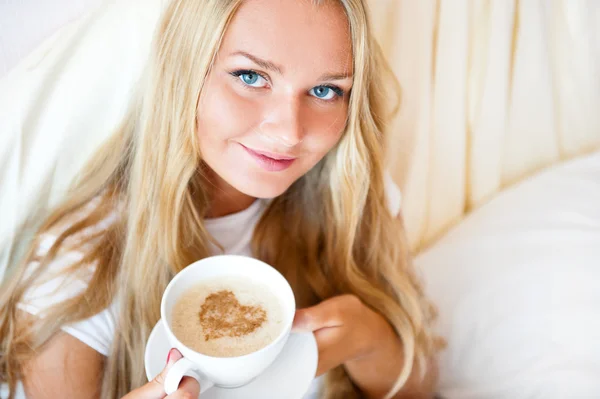 Lachende vrouw drinken een kopje koffie liggend op een bed thuis of hotel. — Stockfoto