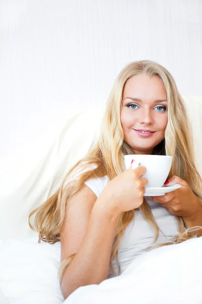 Mujer sonriente bebiendo un café acostada en una cama en casa o en un hotel . —  Fotos de Stock