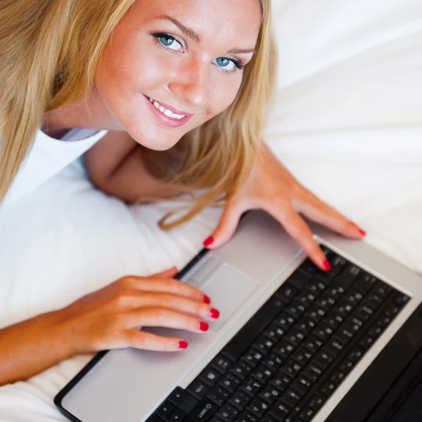 Mujer sonriente usando una computadora portátil mientras está acostada en su cama y comiendo r — Foto de Stock