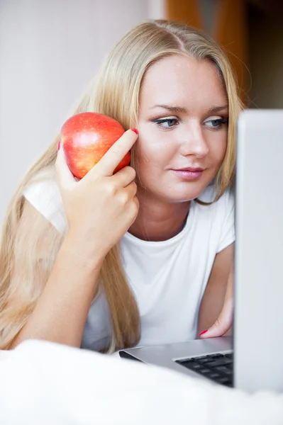 Smiling woman using a laptop while lying on her bed and eating r — Stock Photo, Image