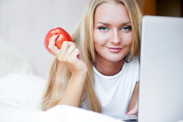Mosolygó nő használ egy laptop, míg hanyatt fekszik az ágyon, és eszik r — Stock Fotó