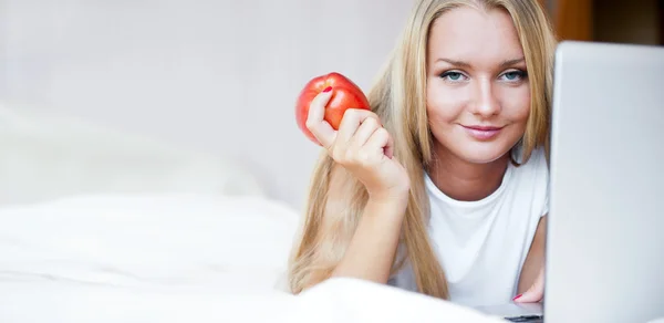 Smiling woman using a laptop while lying on her bed and eating r — Stock Photo, Image