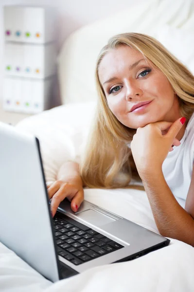Mujer sonriente usando un portátil mientras está acostada en su cama —  Fotos de Stock