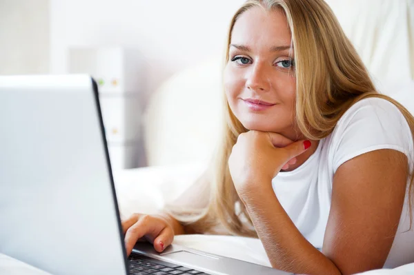 Mulher sorridente usando um laptop enquanto deitada em sua cama — Fotografia de Stock
