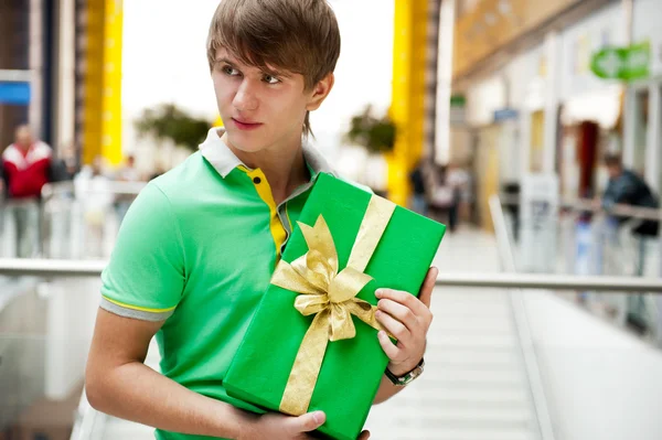 Retrato de hombre joven dentro del centro comercial con caja de regalo sentado — Foto de Stock