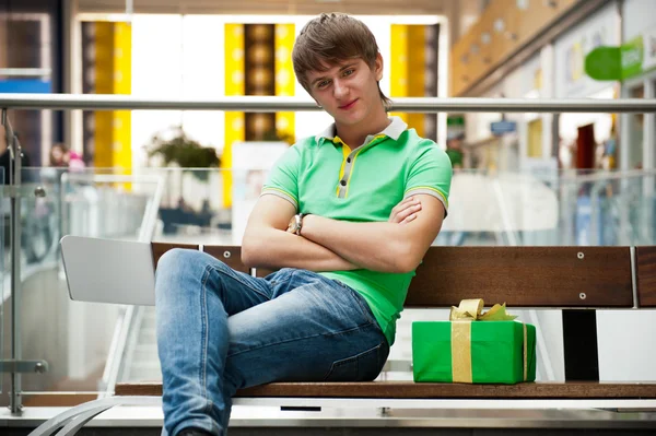 Porträtt av ung man inuti köpcentrum med gåva rutan sitter — Stockfoto