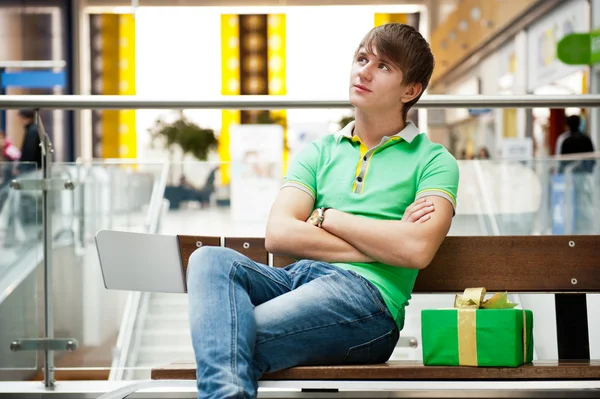 Retrato de hombre joven dentro del centro comercial con caja de regalo sentado — Foto de Stock