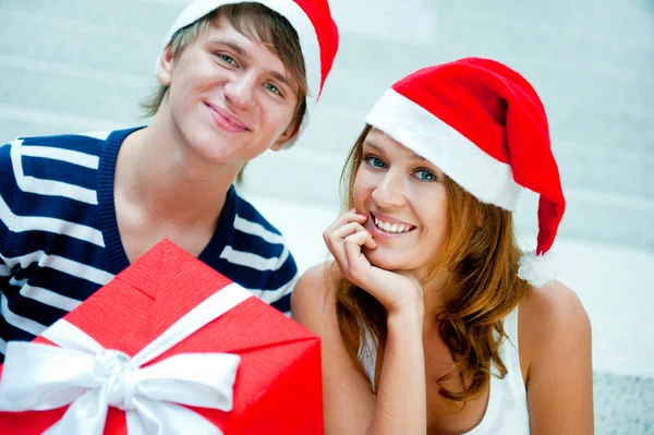 Młoda para szczęśliwy w Boże Narodzenie kapelusze stojąc razem i holdi — Zdjęcie stockowe