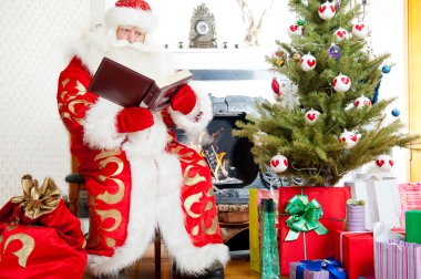 Santa sitting at the Christmas tree, fireplace and reading a boo clipart