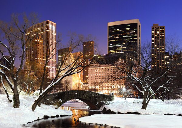 New York City Manhattan Central Park panorama in winter with snow, bridge; freezing lake and skyscrapers at dusk.