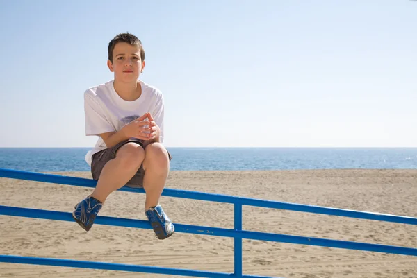 Young boy sitting on a metal railing, beach and sea in the backg — Stock Photo, Image