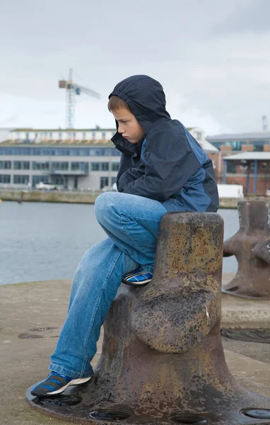Niño infeliz sentado junto al agua en un puerto, lluvia —  Fotos de Stock