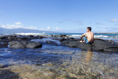 Young man sitting on the lava rocks near ocean in Kapalua Beach, Maui. Hawa clipart