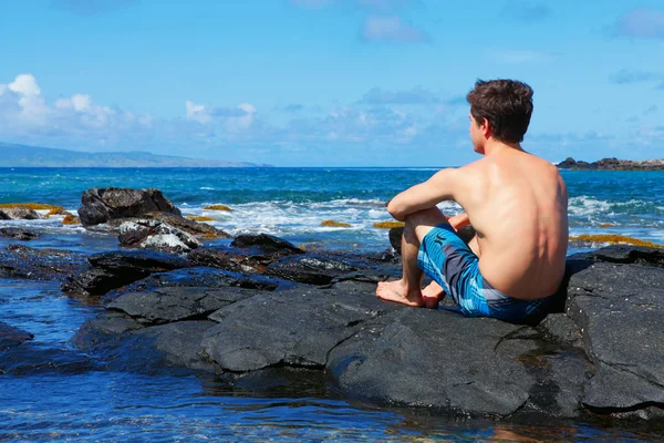 Young man is sitting on the lava rocks near ocean. — Stock Photo, Image