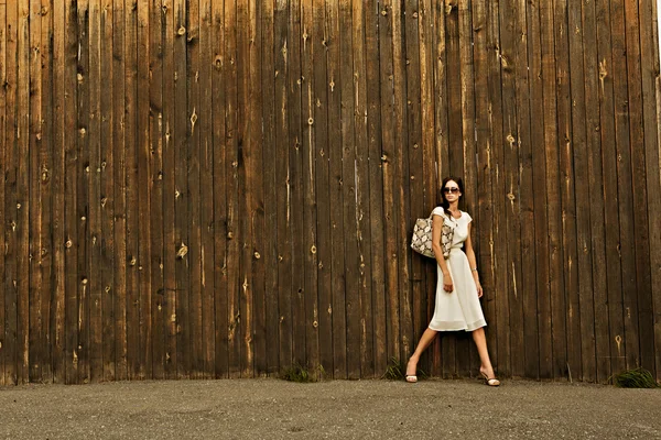 Schöne Mädchen in weißem Kleid und Tasche gegen die Wand mit einer Holzstruktur — Stockfoto