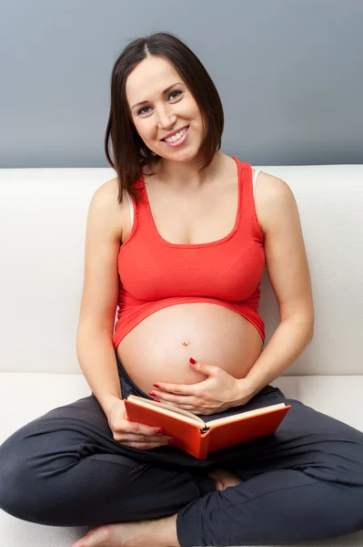 Mujer embarazada sentada en un sofá y sosteniendo el libro —  Fotos de Stock