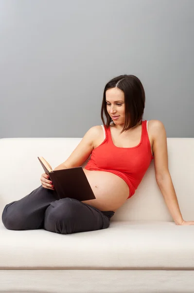 Pregnant woman sitting on sofa and reading book — Stock Photo, Image