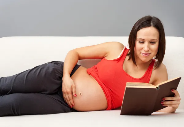 임신한 여자와 소파에 누워 책을 읽고 — 스톡 사진