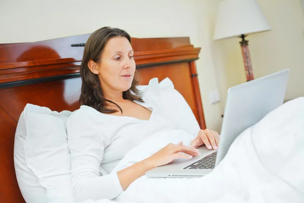 Usmívající se žena pomocí přenosného počítače sedí v posteli — Stock fotografie