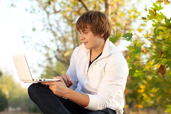 年轻男子坐在一个长椅上有一台笔记本电脑，秋天 — 图库照片