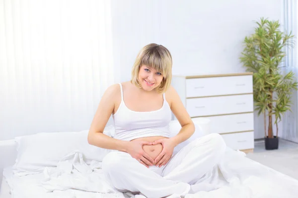 Kobieta siedzi na białej kanapie w domu i co serca na jej brzuch w ciąży — Zdjęcie stockowe