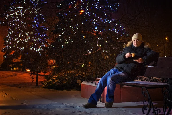 A young man sits on a bench and drink coffee at Christmas, snowfall — Stock Photo, Image