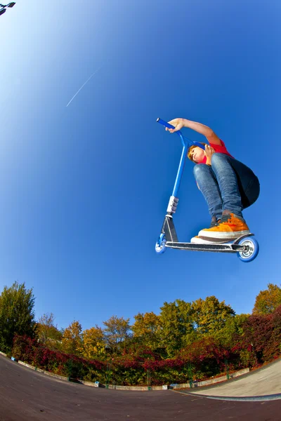 Ragazzo che salta con il suo scooter allo skate park sotto blu chiaro — Foto Stock