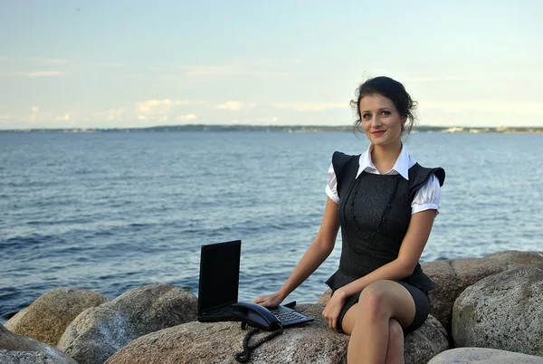 Oficina junto al mar: la chica en las rocas con un ordenador portátil y un teléfono de escritorio — Foto de Stock