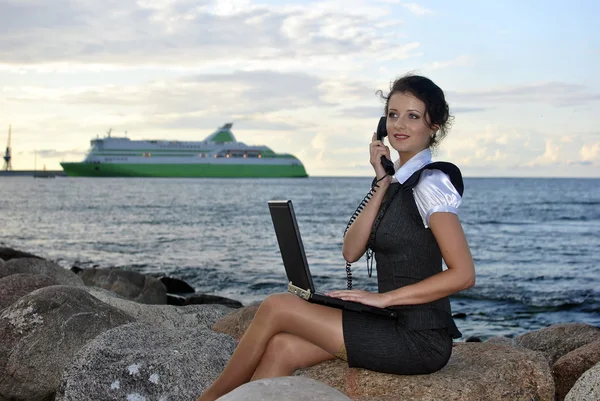 Ufficio in riva al mare: la ragazza sugli scogli con un computer portatile e un telefono desktop — Foto Stock
