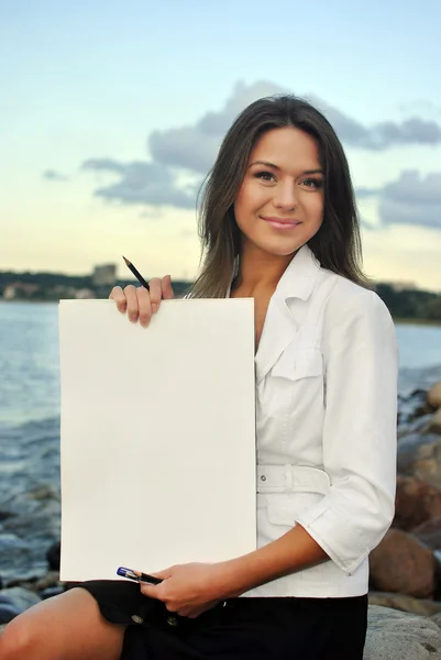 Menina bonita em uma jaqueta branca segurando papel em branco. mar no fundo — Fotografia de Stock