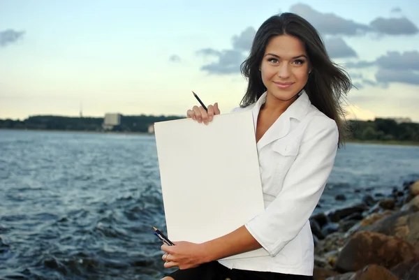 Schönes Mädchen in weißem Mantel mit leerem Papier. Meer auf dem Hintergrund — Stockfoto