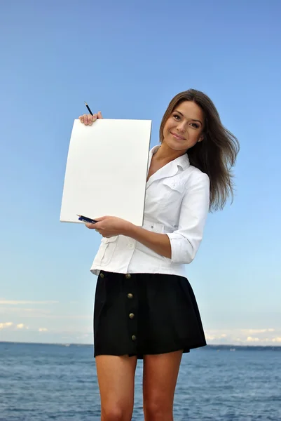 Hermosa chica con una chaqueta blanca sosteniendo papel en blanco. mar en el fondo —  Fotos de Stock