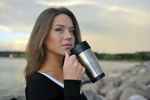 Hermosa chica de pie en la playa con una taza de café y teléfono celular — Foto de Stock